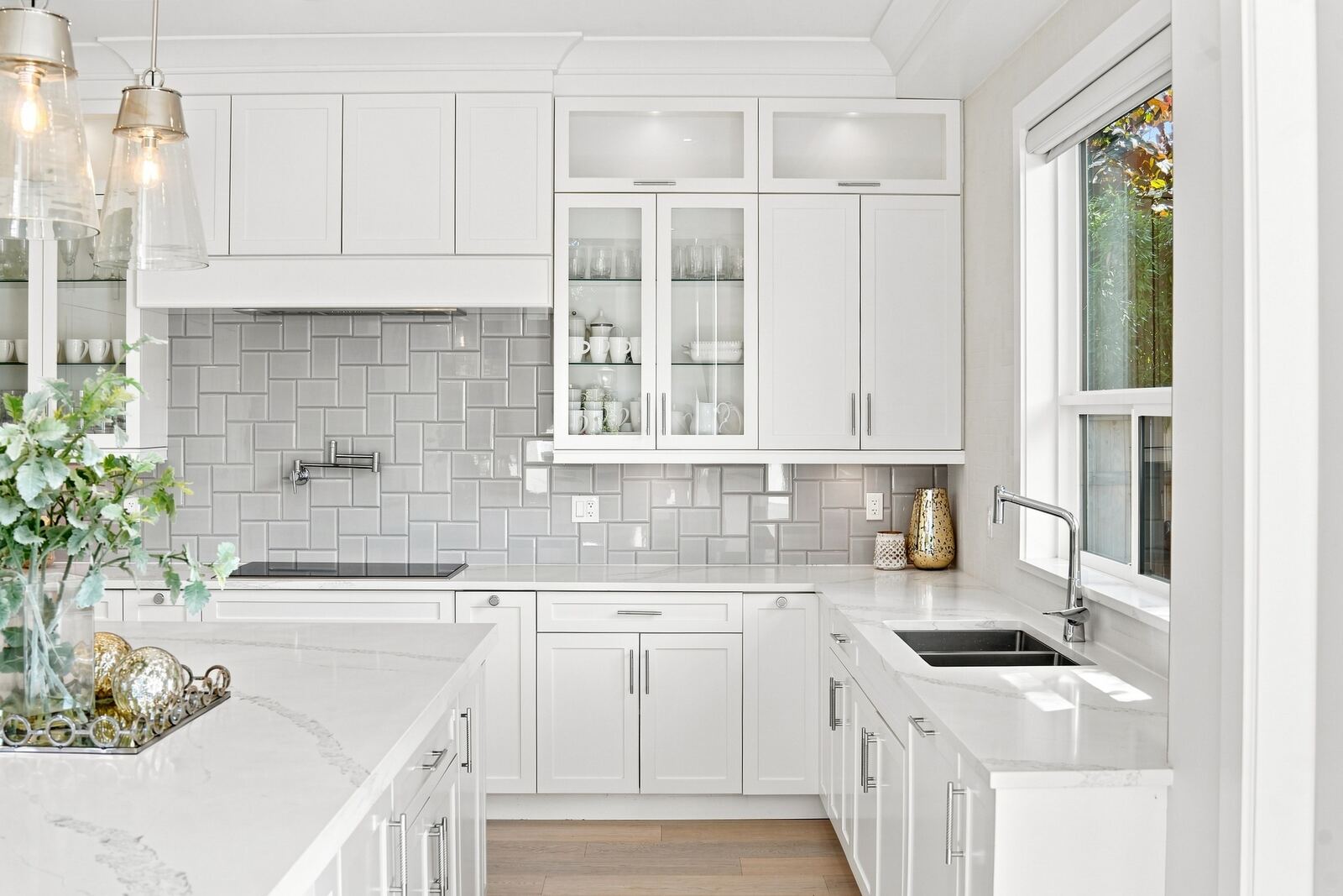 Bright white kitchen with quartz countertops, glass-front cabinets, grey tile backsplash and a window over the sink