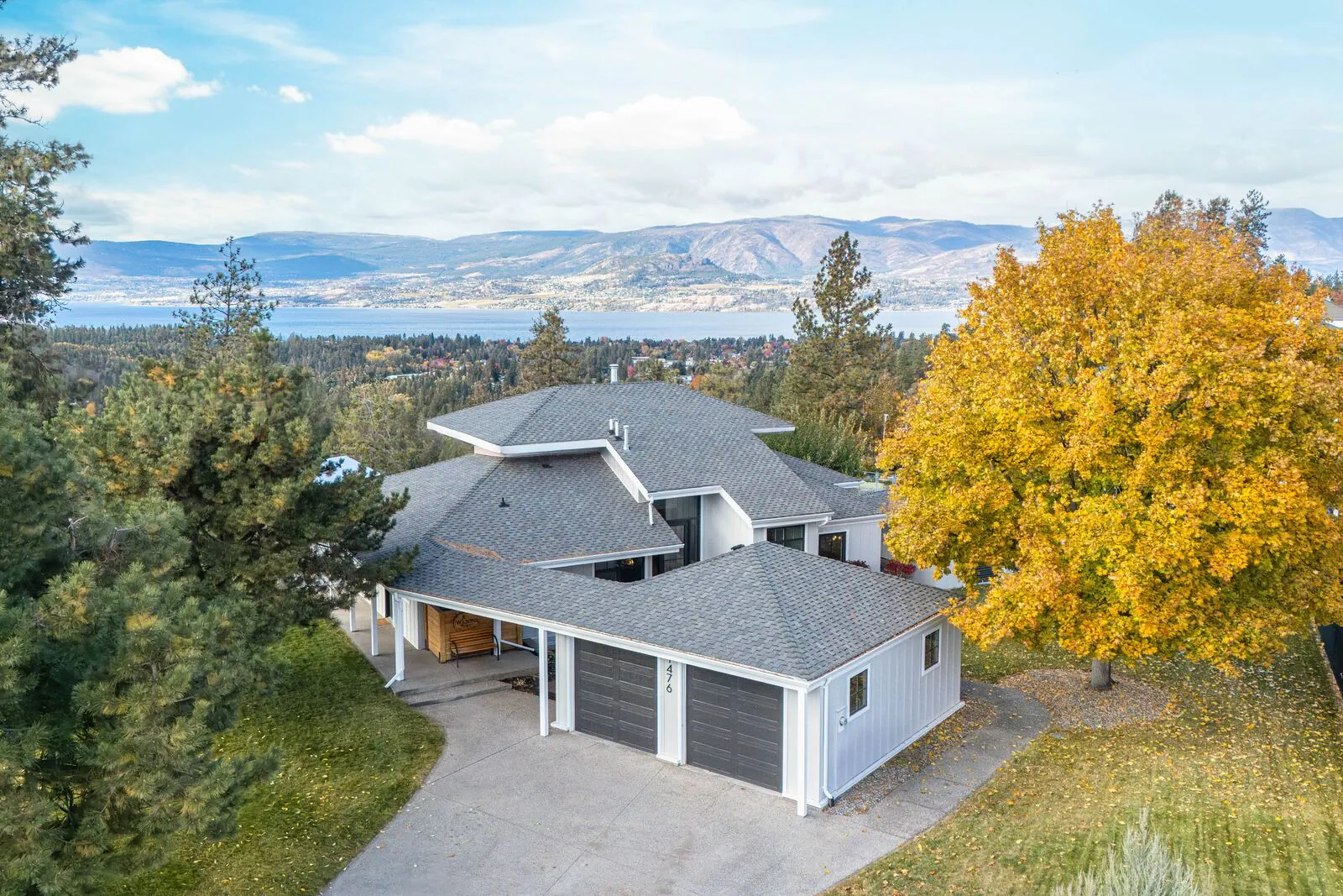 Aerial drone photo of a grey-roofed home with a triple garage, autumn trees and Okanagan Lake with mountains beyond