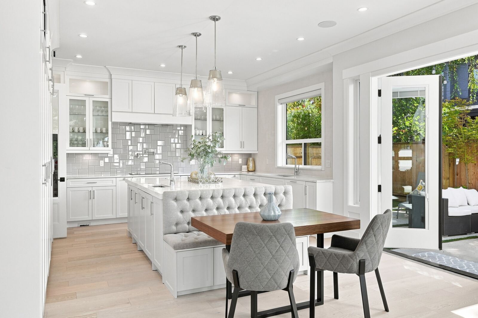 Open-concept white kitchen with a tufted banquette dining nook, pendant lights and a patio door