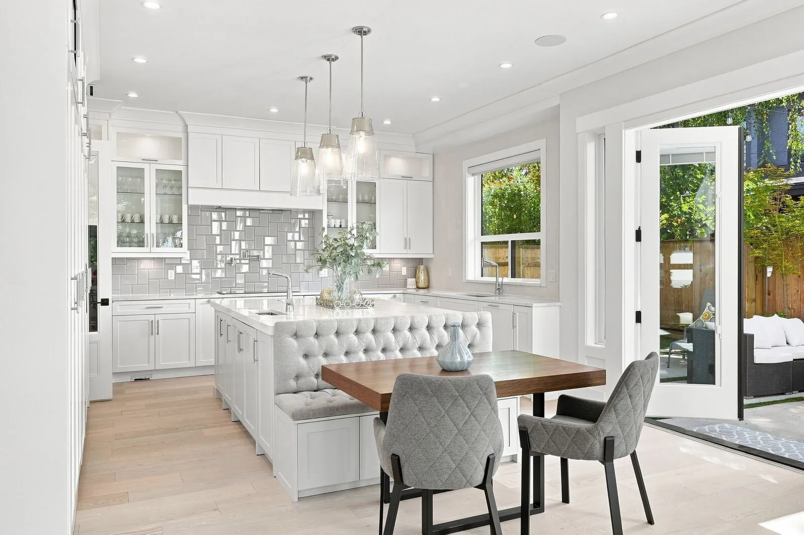 Open-concept white kitchen with a tufted banquette dining nook, pendant lights and a patio door