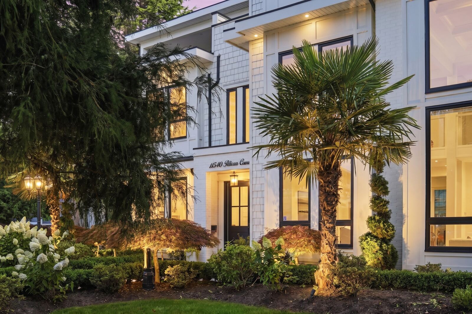 White home entrance at evening with warm window glow, a palm tree and landscape lighting