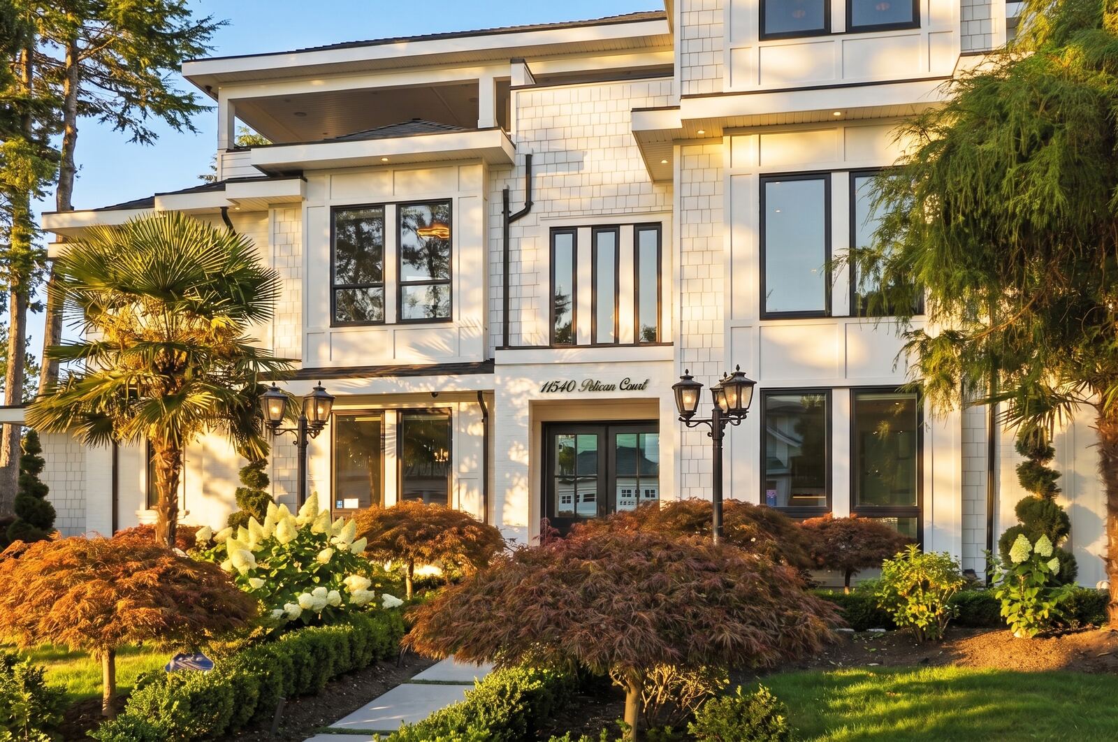 White two-storey home entrance in golden-hour light with Japanese maples and lantern-style lamps